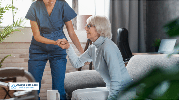 A caregiver helping a senior woman with mobility, gently assisting her while standing up, with the Always Best Care logo in the bottom-left corner.