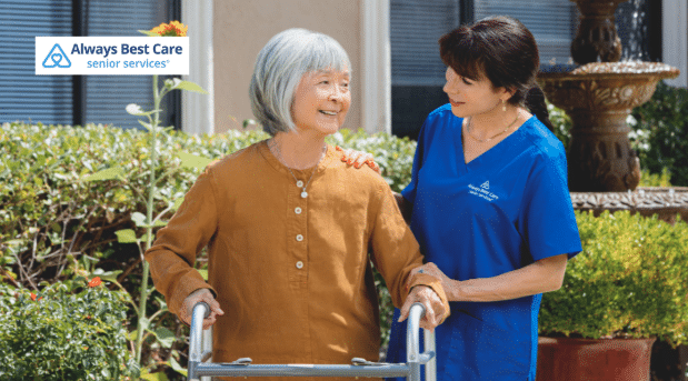 A caregiver helping a senior woman with mobility, offering her a steady hand while she stands, with the Always Best Care logo in the bottom-left corner.