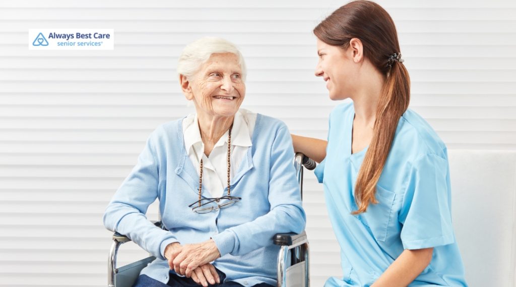 A caregiver in blue scrubs is smiling at a senior woman in a wheelchair. The woman is dressed in a light blue cardigan and looks happy and comfortable, indicating a supportive, caring environment where the elderly feel valued and attended to.