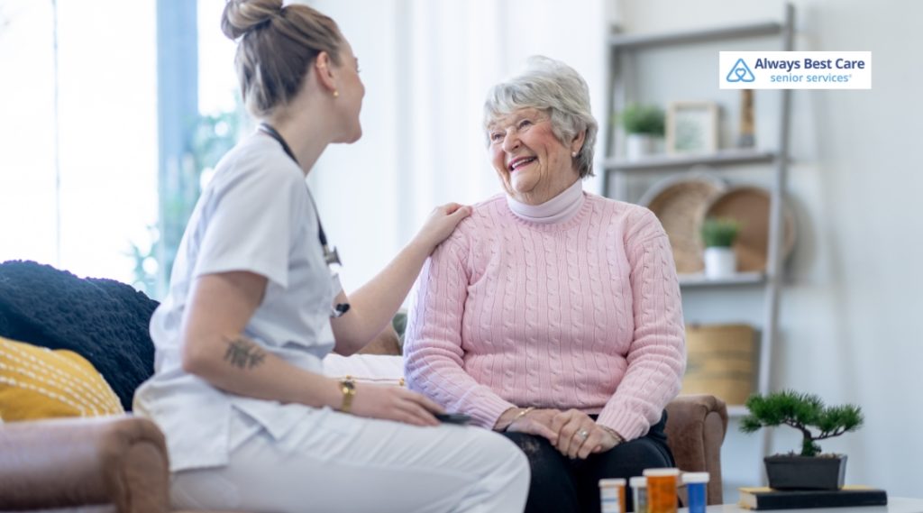 A caregiver sits next to an elderly woman, smiling and interacting with her in a cozy living room, with medication bottles visible on the table.