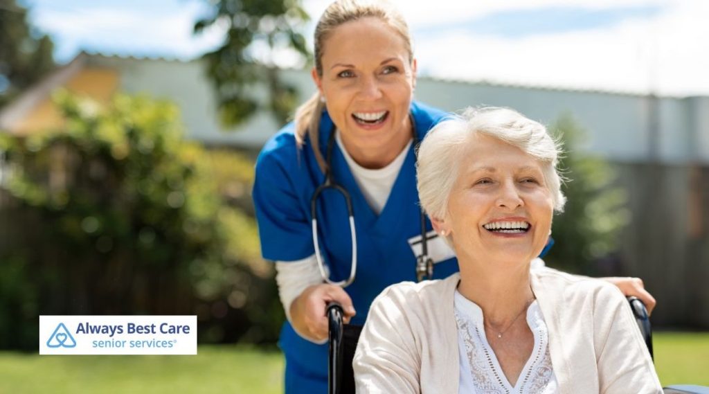 Happy older woman in a wheelchair laughing outdoors while a nurse in blue scrubs stands behind her, both smiling, with the Always Best Care senior services logo visible.