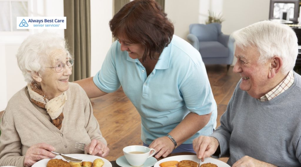 Friendly caregiver chats with a senior couple during lunch, highlighting positive caregiver relationships and daily support.