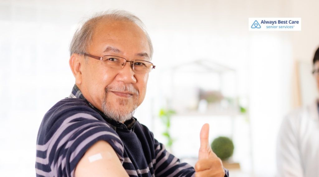 A senior man in a striped sweater smiling and giving a thumbs-up after receiving a vaccine shot, with a band-aid on his arm.