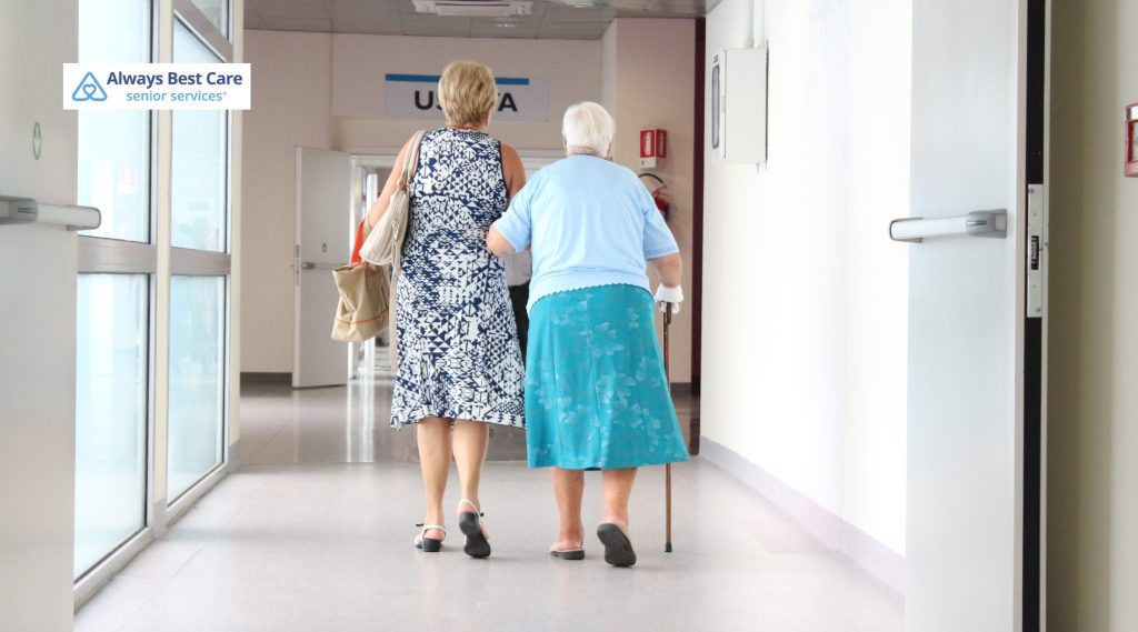 A caregiver walking alongside a senior woman down a hospital hallway, with the "Always Best Care" logo in the corner.