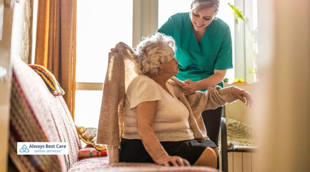 A caregiver helps an elderly woman put on a cardigan, smiling warmly in a sunlit living room, representing compassionate in-home support.