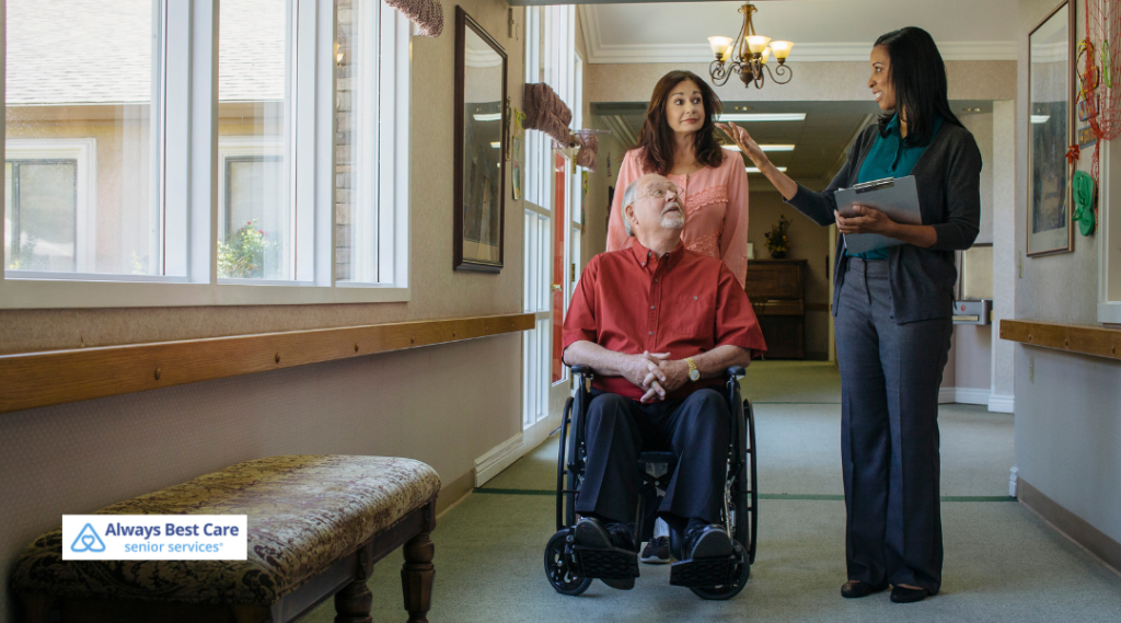Two caregivers walk alongside an elderly man in a wheelchair through a care facility hallway, engaging in friendly conversation while showing him around.