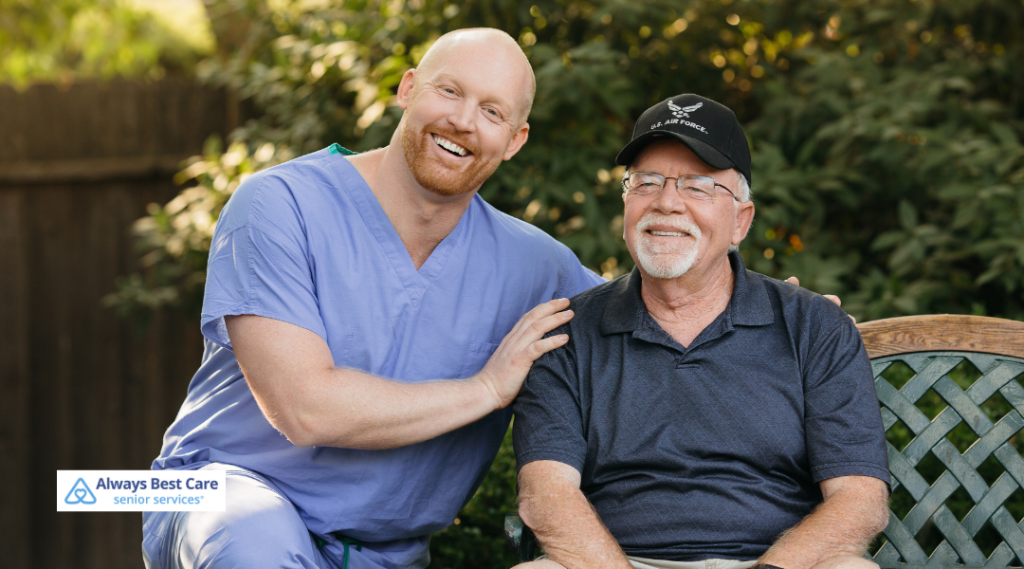 A cheerful caregiver in blue scrubs smiles beside an older man wearing glasses and a U.S. Air Force hat, as they enjoy a sunny day sitting on a garden bench.