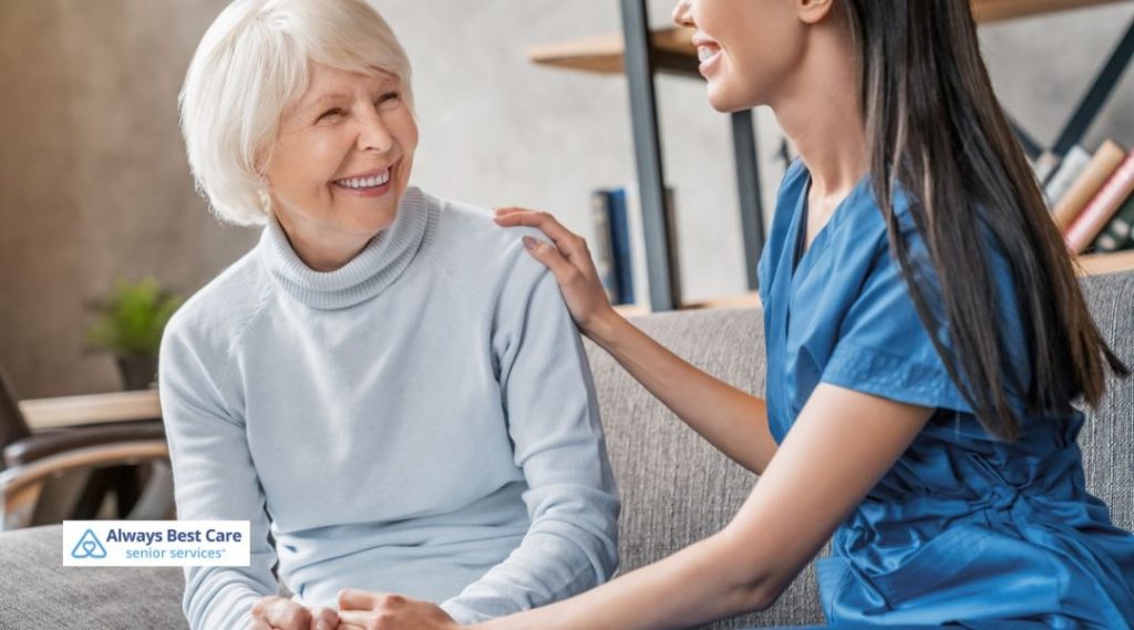 A smiling caregiver sits beside a senior woman on a couch, offering comfort and emotional support through friendly conversation.