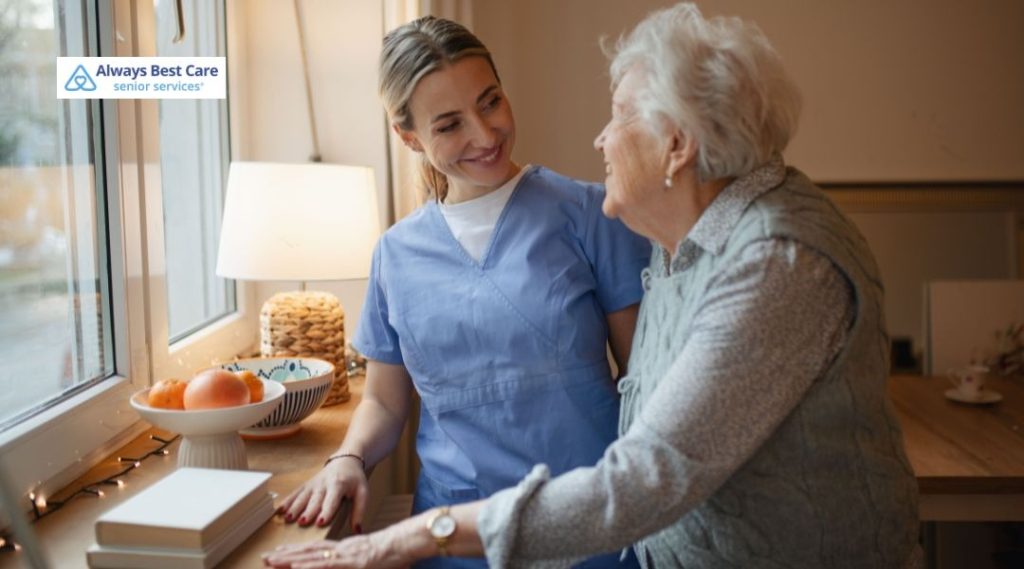 A caregiver and a senior woman share a moment by a window, with the caregiver offering a comforting touch and both smiling. This highlights the emotional connection fostered through Always Best Care of Westerville’s services.