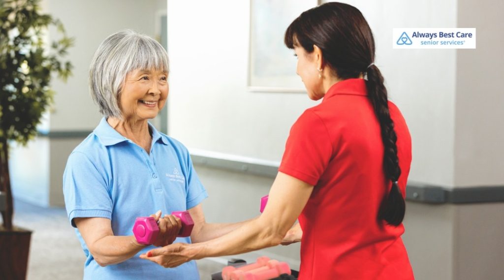 Always Best Care caregiver helping a senior woman perform arm exercises with a pink hand weight.