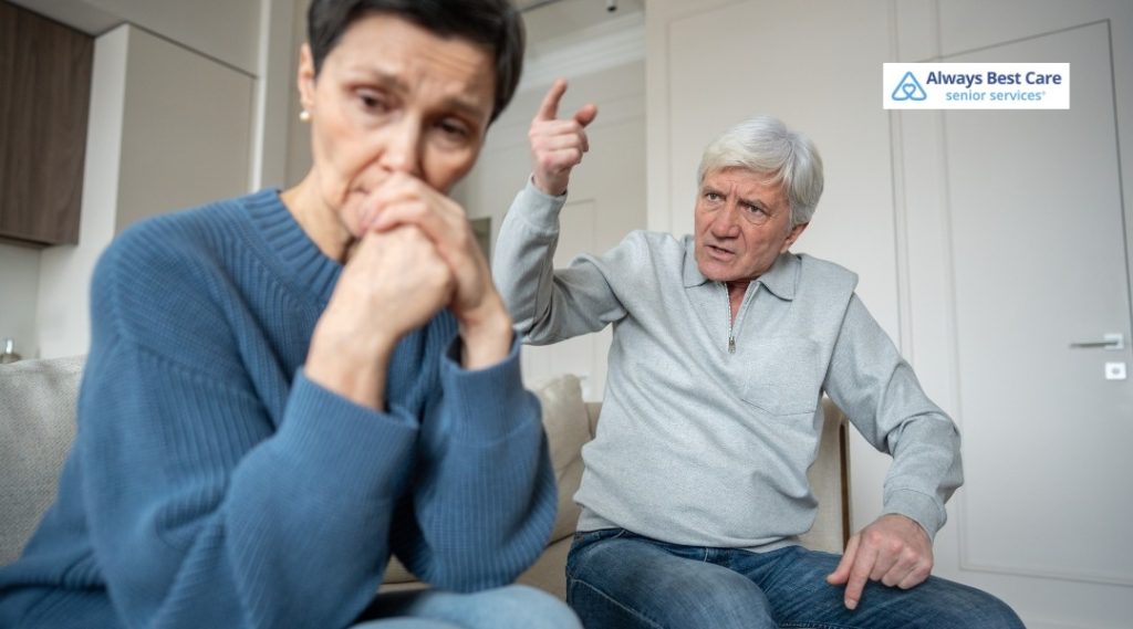 Older man on a sofa angrily gesturing toward a distressed person sitting nearby, highlighting a tense moment at home, Always Best Care logo in corner.