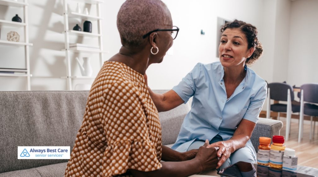 A compassionate caregiver is sitting beside an elderly woman, smiling and holding her hands while discussing medication. The caregiver is wearing a light blue uniform and the woman is dressed in a polka-dot blouse. Various pill bottles are seen in front of them, indicating medication management.