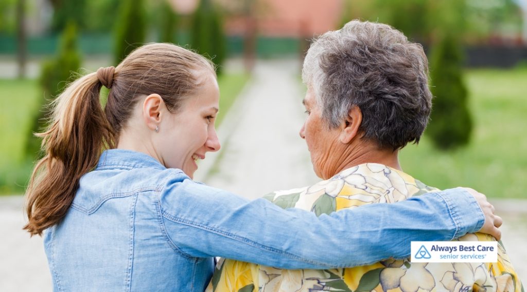 A caregiver is walking with an elderly woman, offering her support as they stroll outdoors in a calm, green environment.