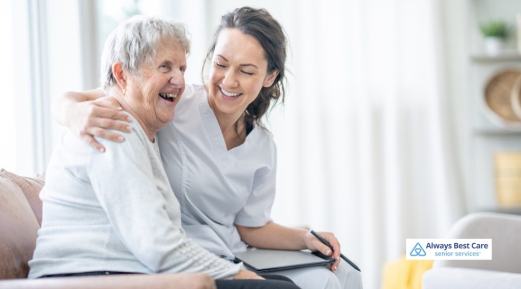 A caregiver and an elderly woman share a joyful moment together in a bright and airy room, with the caregiver holding a tablet and offering assistance.