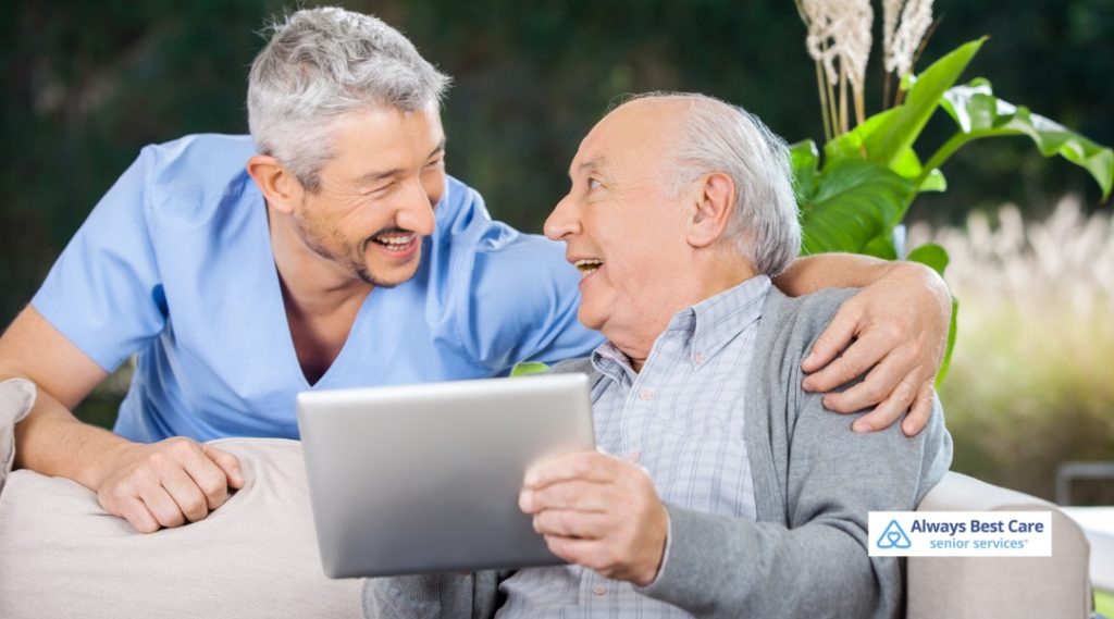 A caregiver in blue scrubs shares a joyful moment with an elderly man, both laughing together as the man holds a tablet. They sit together on a comfortable sofa in a peaceful, natural setting.