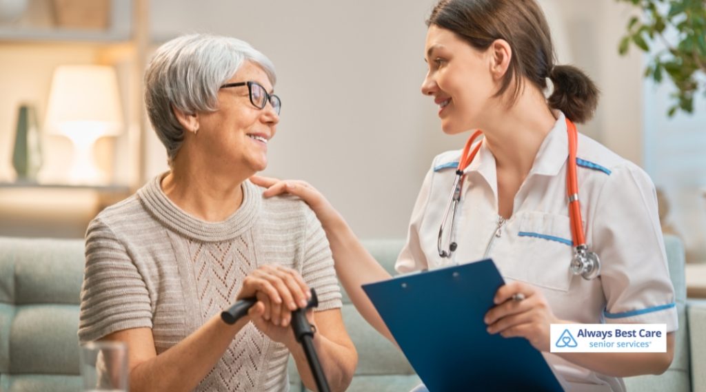 A healthcare professional in a white uniform interacts with an elderly woman holding a cane. The professional is offering guidance, and the woman smiles in a comforting, home-like environment.