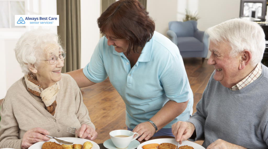 A caregiver and a senior couple smiling and enjoying a meal together at the table. The caregiver is standing behind the woman, offering support. BrightStar Care logo in the corner.
