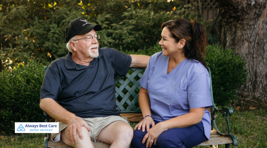 A senior man in a baseball cap and a caregiver sitting together on a park bench, both smiling and enjoying each other’s company. BrightStar Care logo in the corner.