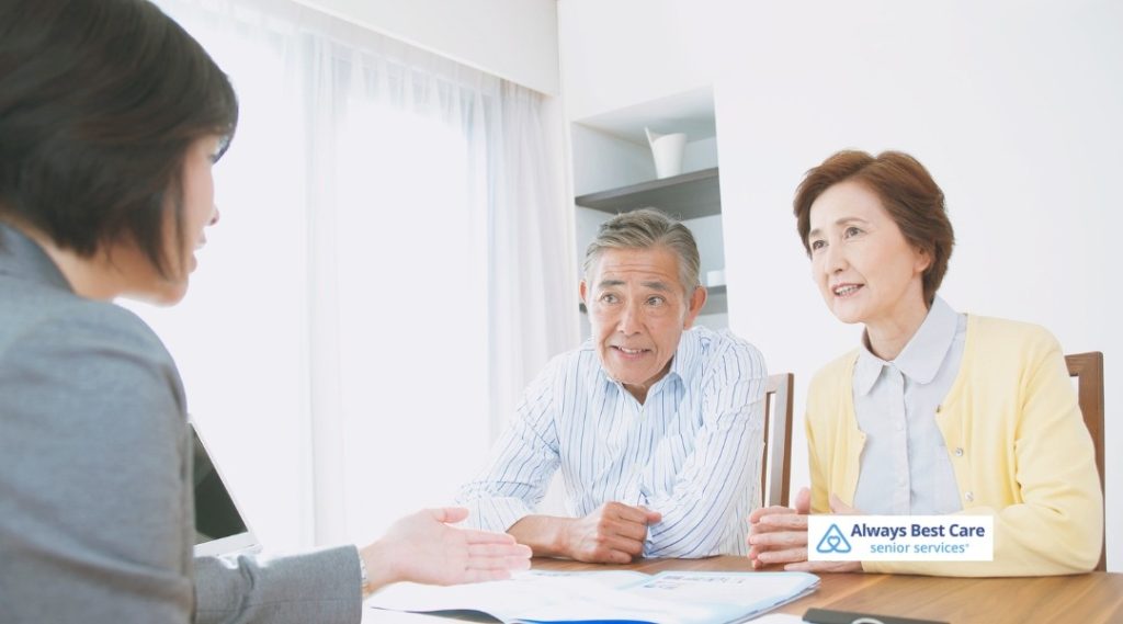 A senior couple attentively listens to a professional during a meeting at home. The couple looks engaged and relaxed, and the Always Best Care logo is displayed in the lower right corner.