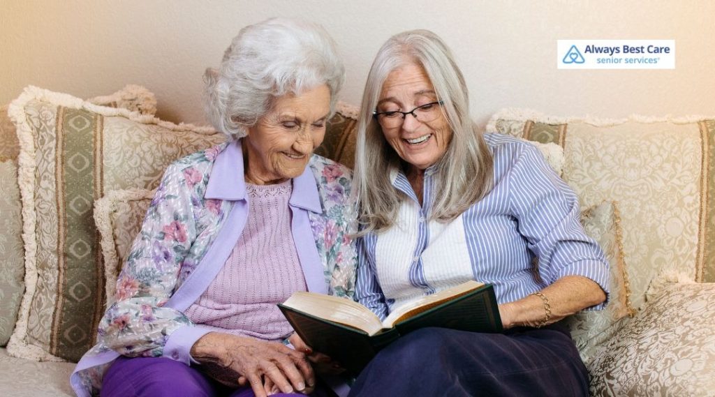 Two older women sitting close together on a sofa, reading a book and sharing a quiet moment, with the Always Best Care senior services logo in the corner.