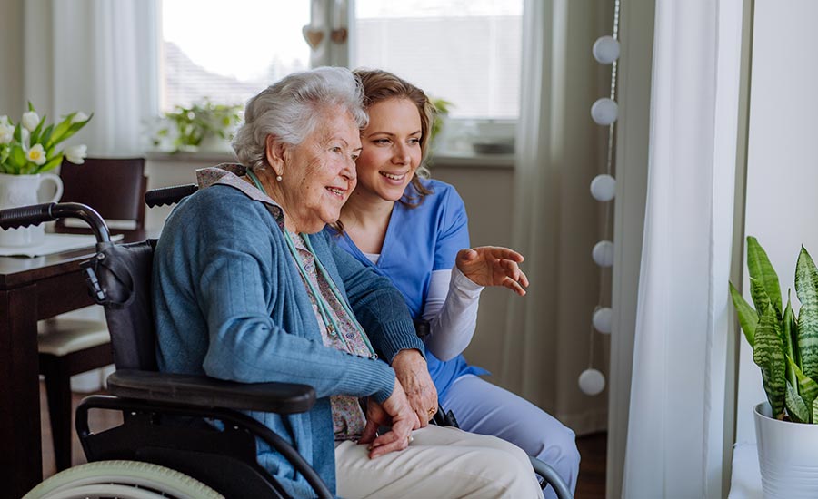 An elderly woman smiles with her nurse while looking out the window​
